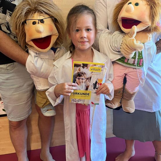 Ein junges Mädchen hält ein Buch in einer Laborkittel, flankiert von zwei Puppen in Laborkitteln, posiert drinnen.