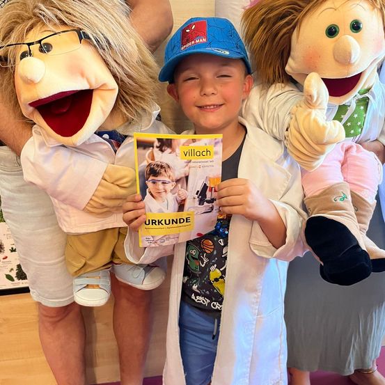 Ein junger Junge trägt einen weißen Laborkittel und hält ein buntes Magazin mit dem Titel 'Urkunde', während zwei Puppen in Kostümen neben ihm stehen, eine davon zeigt einen Daumen hoch.
