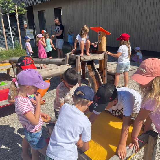 Eine Gruppe von Kindern spielt in einem Outdoor-Aktivitätsbereich. Einige sind in einem hölzernen Sandkasten, während andere einen hölzernen Wasserbrunnen benutzen. Erwachsene beaufsichtigen im Hintergrund.