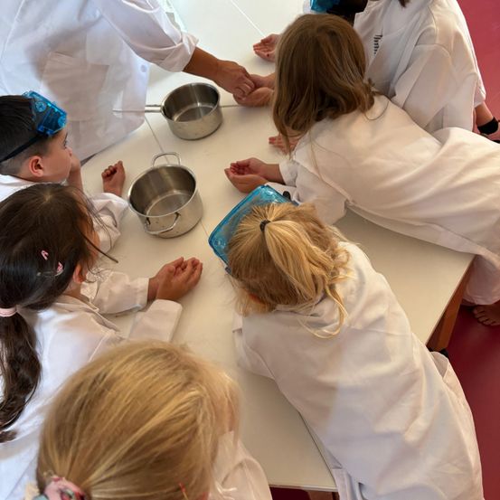 Kinder in weißen Laborkitteln versammeln sich um einen Tisch mit Töpfen und nehmen an einem wissenschaftlichen Experiment teil.