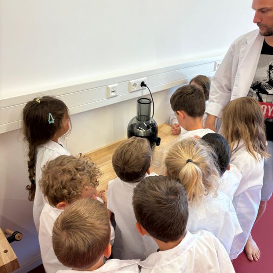 Kinder in weißen Laborkittel versammeln sich um einen Mixer auf einem Holztisch, angeführt von einem Mann in einem Laborkittel. Sie scheinen an einem wissenschaftlichen Experiment teilzunehmen.
