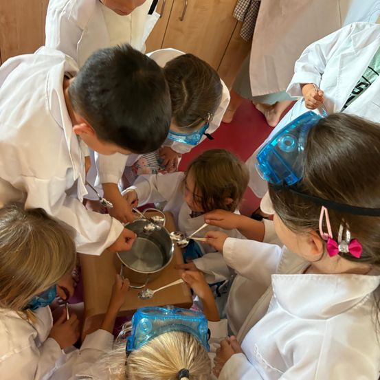 Mehrere Kinder in weißen Laborkittel und Schutzbrillen sind um einen Tisch versammelt und rühren und messen Zutaten in einem Topf.