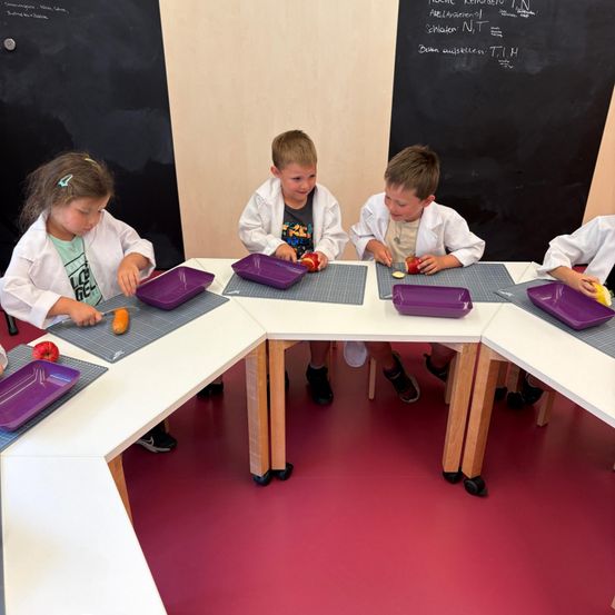 Vier Kinder in Laborkittel sitzen um einen Tisch herum, jeder mit einem lila Teller und schneidet Gemüse. Hinter ihnen steht eine Tafel mit einigen mathematischen Gleichungen und Formeln.