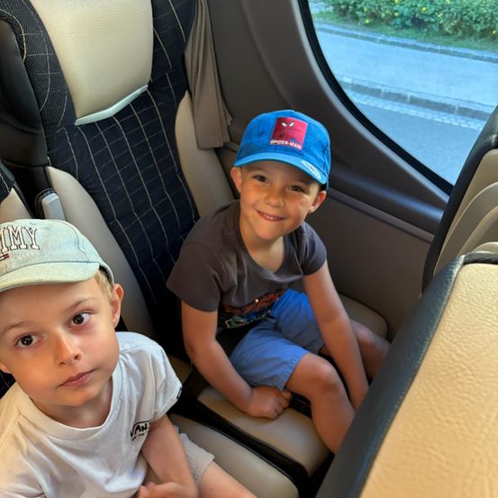 Zwei Jungen sitzen in einem Fahrzeug, einer mit einem Spider-Man-Hut und der andere mit einem Jimmy-Hut. Sie schauen aus dem Fenster auf eine Straße.