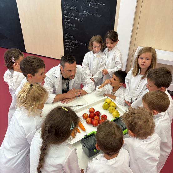 Eine Gruppe von Kindern in weißen Laborkitteln versammelt sich um einen Tisch mit Obst und Gemüse. Ein erwachsener Mann steht vor dem Tisch und erklärt den Kindern etwas. Hinter ihnen befindet sich eine Tafel mit ein paar darauf geschriebenen Texten.