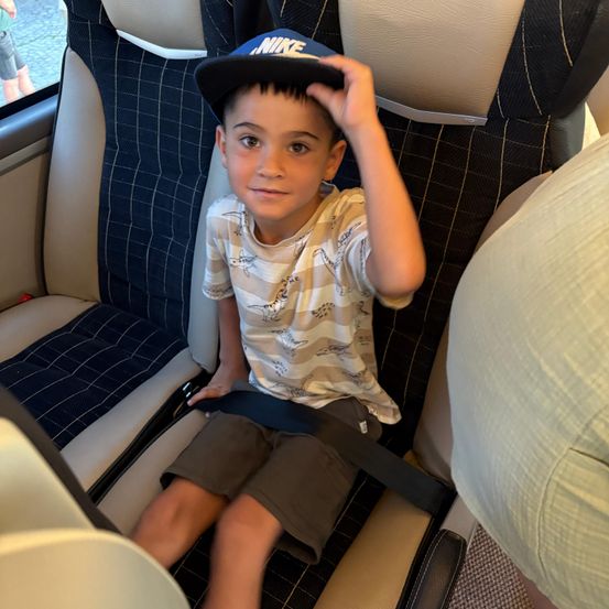 Ein junger Junge mit einer Nike-Kappe und einem T-Shirt mit Dinosaurier-Aufdruck sitzt in einem Fahrzeug. Er hat einen Sicherheitsgurt angelegt und lächelt in die Kamera.