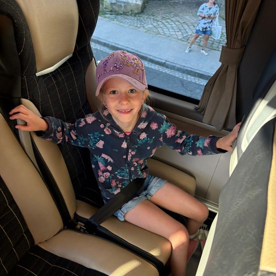 Ein kleines Mädchen mit einem Blumenhemd und einer Kappe sitzt in einem Bus, lächelt und schaut aus dem Fenster, wo eine Frau auf einem Kopfsteinpflasterweg geht.