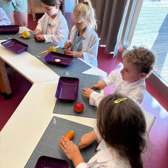 Mehrere Kinder in Laborkitteln sitzen um einen Tisch herum, jeder mit einem lila Tablett und Schneidwerkzeugen, und bereitet verschiedene Früchte zu. Ein Junge schneidet einen Apfel und ein Mädchen schneidet eine Karotte. Hinter ihnen ist eine Glastür mit einer Holzterrasse.
