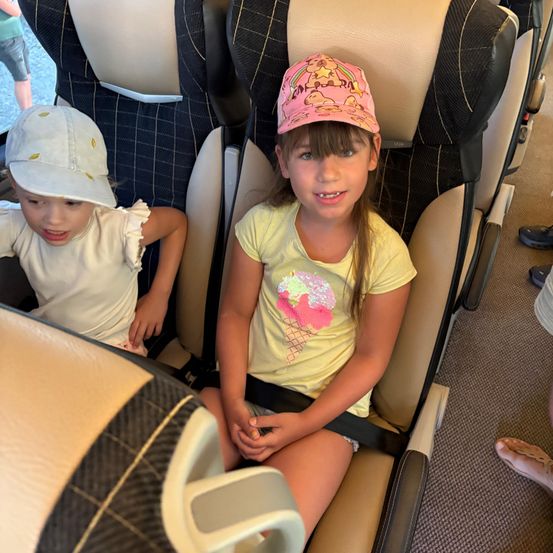 Zwei junge Mädchen sitzen in Flugzeugsitzen, eines trägt ein gelbes Shirt mit Eiscreme-Design und eine rosa Mütze. Das andere Mädchen hat eine weiße Mütze und ein weißes Shirt. Hinter ihnen ist der Fuß einer Person und ein Kinderwagen zu sehen.