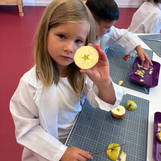 Ein junges Mädchen in einer weißen Laborkittel hält einen geschnittenen Apfel in der Hand und steht an einem Tisch mit Schneidematten. Ein anderes Kind, ebenfalls in einem Laborkittel, ist beim Schneiden von Äpfeln zu sehen.