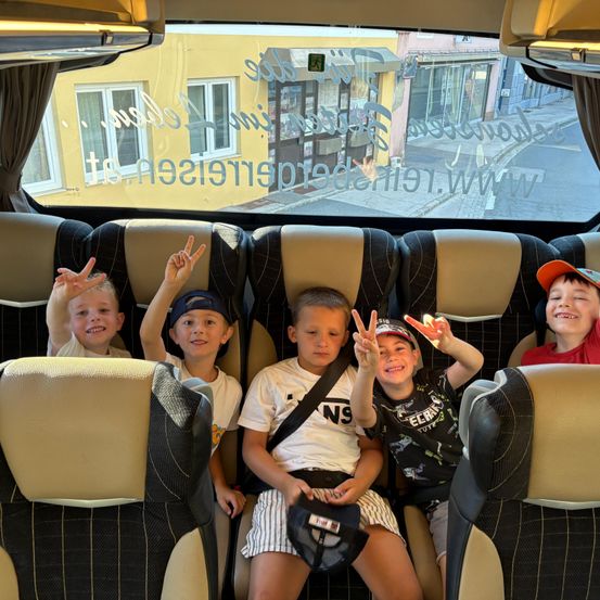 Fünf Jungen sitzen in einem Bus mit einem gelben Gebäude im Hintergrund. Vier Jungen machen Friedenszeichen. Der Junge in der Mitte trägt einen Sicherheitsgurt.