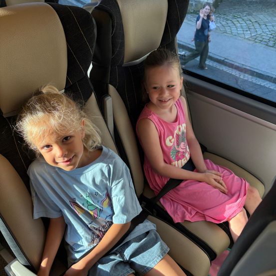Zwei junge Mädchen sitzen in einem Bus, lächeln und schauen aus dem Fenster. Das Mädchen auf der linken Seite trägt ein blaues Shirt mit einem Einhorn-Design. Das Mädchen auf der rechten Seite trägt ein rosa Kleid. Hinter ihnen laufen eine Frau und ein Kind auf dem Bürgersteig.
