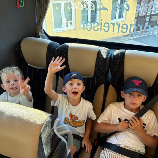 Drei junge Jungen sitzen in einem Bus. Der Junge in der Mitte winkt enthusiastisch. Der Junge auf der rechten Seite hält seinen Sicherheitsgurt, und der Junge auf der linken Seite macht ein Friedenszeichen.