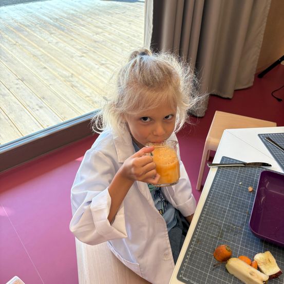 Ein kleines Mädchen in einer Laborkittel trinkt Orangensaft an einem Tisch mit geschnittenen Früchten. Der Raum hat eine Glaswand mit Blick auf eine Holzterrasse.