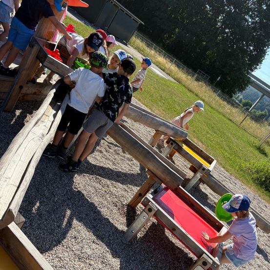Eine Gruppe von Kindern spielt in einem hölzernen Spielbereich mit verschiedenen Holzstrukturen und rotem Sand. Einige graben, während andere zuschauen.