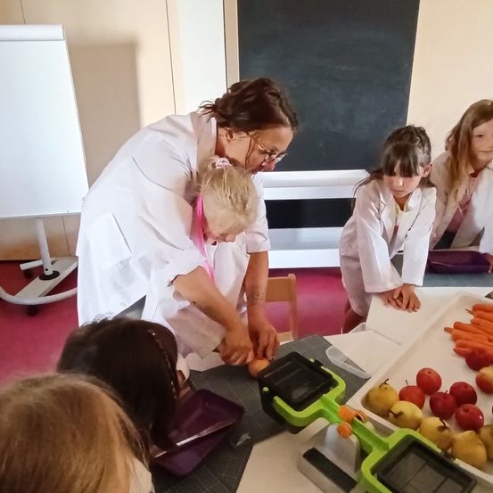 Eine Frau in einem weißen Laborkittel unterrichtet junge Mädchen, wie man einen Apfel schält. Die Mädchen tragen Laborkittel und arbeiten gemeinsam an einem Tisch mit Äpfeln und Karotten.