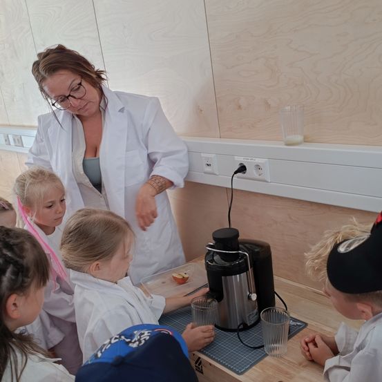 Eine Frau in einem weißen Laborkittel zeigt einer Gruppe von Kindern, wie man einen Entsafter benutzt. Die Kinder sitzen an einem Holztisch mit Gläsern und einer Tablett mit Äpfeln. Die Frau demonstriert, während die Kinder aufmerksam zuschauen.