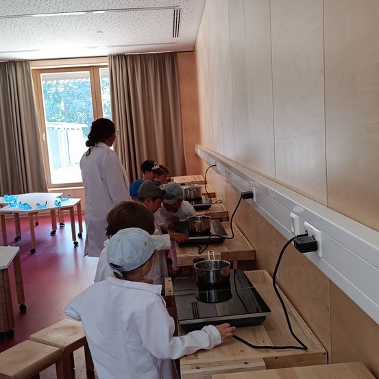 Eine Gruppe von Kindern kocht in einem Klassenzimmer. Sie tragen weiße Mäntel und Hüte. Die Lehrerin steht in der Nähe. Das Klassenzimmer hat Holzwände und -böden. Es gibt Tische und Hocker im Raum.