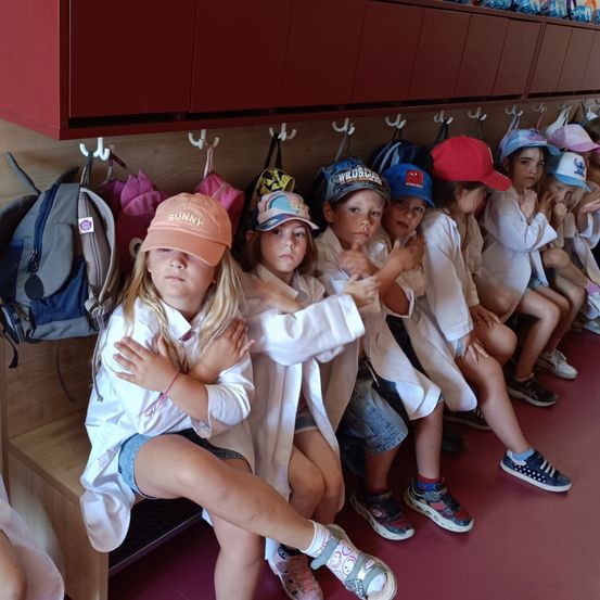 Eine Gruppe von Kindern in Laborkittel sitzen auf einer Bank in einem Raum. Jeder hat einen Rucksack, der an einem Haken über ihnen hängt. Sie wirken konzentriert, einige machen Gesten.
