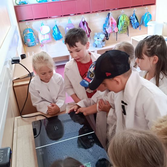 Eine Gruppe von Kindern in Laborkitteln versammelt sich um einen Touchscreen in einem Klassenzimmer. Sie betrachten etwas auf dem Bildschirm. An der Wand dahinter hängen Taschen.