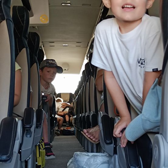 In einem Fahrzeug sitzt ein junger Junge in der mittleren Reihe, während zwei andere Jungen hinten sitzen. Ein Junge schläft mit dem Kopf auf einem Sitz, und mehrere Taschen liegen auf dem Boden.