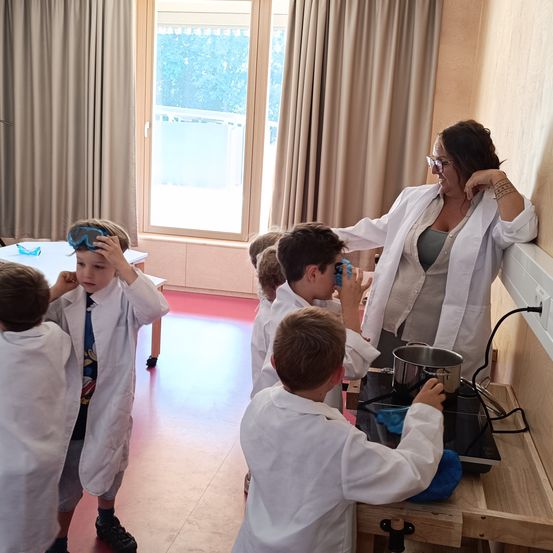 Eine Frau in einem Laborkittel steht hinter Kindern in Laborkitteln und hilft ihnen bei einem wissenschaftlichen Experiment in einem Raum mit Fenster und Herd.