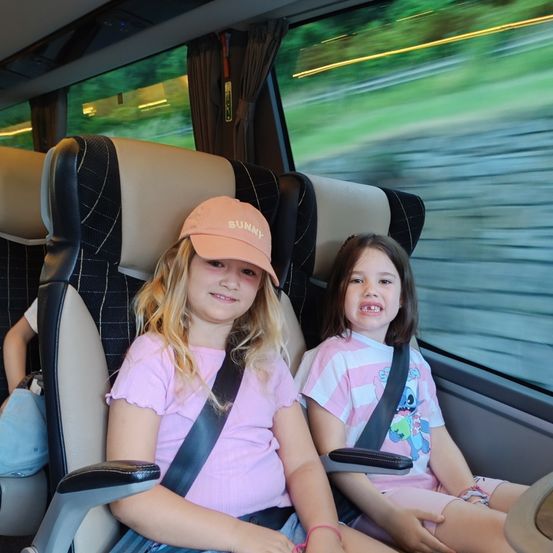 Zwei junge Mädchen in rosa T-Shirts sitzen nebeneinander in einem Bus. Sie lächeln beide. Hinter ihnen zeigt ein Fenster einen verschwommenen Hintergrund einer Straße.