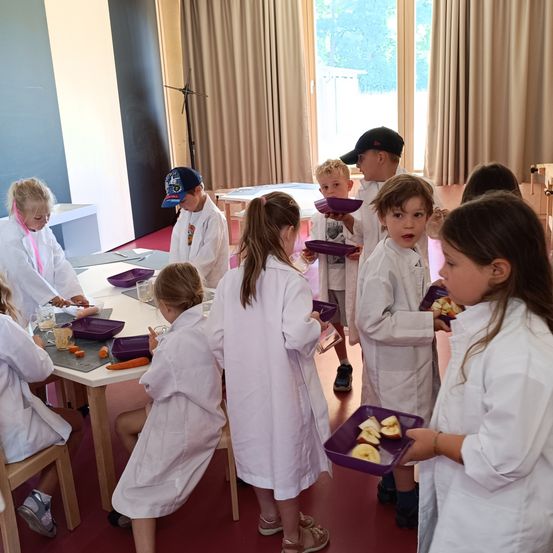 Eine Gruppe von Kindern in weißen Laborkitteln bereitet Essen in einem Klassenzimmer zu. Einige halten lila Teller mit Essen, während andere zuschauen. Tische mit Besteck und Gemüse sind in der Nähe.