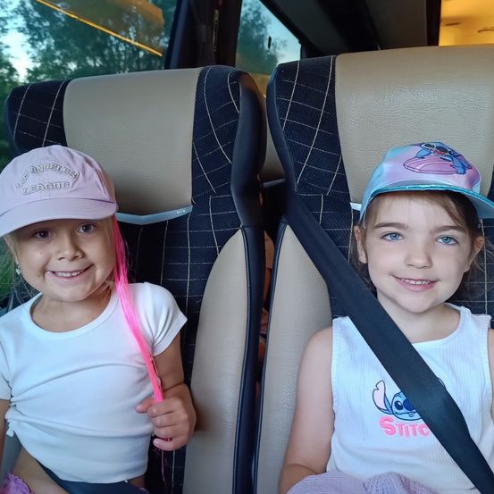 Zwei kleine Mädchen sitzen auf einem Bussitz mit Sicherheitsgurten. Eine trägt eine rosafarbene Baseball-Kappe mit rosa Haaren. Die andere trägt ein weißes Tanktop mit einem Stitch-Charakter darauf.