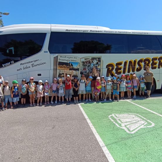 Eine Gruppe von Kindern posiert vor einem Bus mit den Worten 'Reinsberg Reisen' an der Seite. Sie tragen Mützen und Turnschuhe. Der Bus hat ein großes Fenster an der Seite.