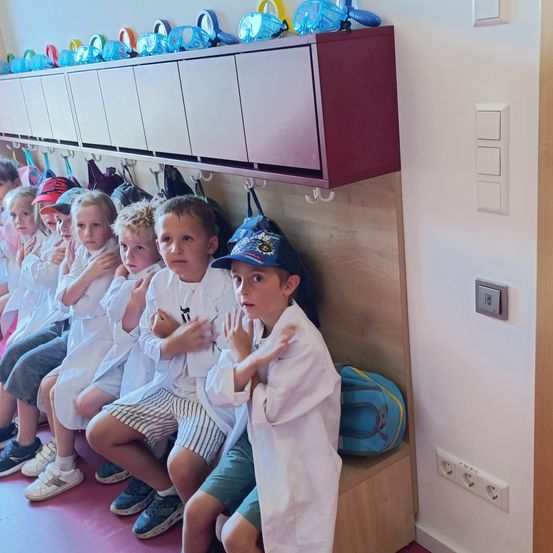 Eine Gruppe von Kindern in weißen Laborkittel sitzen auf einer Bank in einem Raum, mit mehreren farbigen Taucherbrillen, die über ihnen an einem Regal hängen.