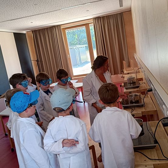 Eine Gruppe von Kindern in weißen Laborkittel und Schutzbrillen beobachtet eine Frau in einem Laborkittel, die ein wissenschaftliches Experiment mit Töpfen auf einem Herd durchführt.