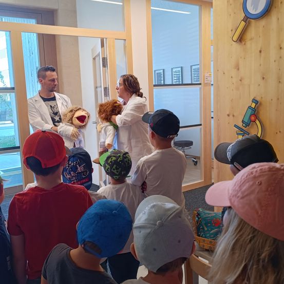 Eine Gruppe von Kindern und zwei Erwachsenen sind in einem Raum mit einer Holzwand und einer Glastür. Die Erwachsenen halten Puppen und scheinen etwas zu erklären.