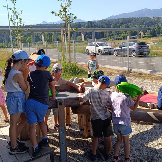 Kinder spielen in einem Sandkasten draußen, tragen Mützen und Turnschuhe. In der Nähe ist eine Straße mit einem Zaun abgegrenzt, und im Hintergrund befindet sich eine Brücke. Zwei Autos sind am Straßenrand geparkt.