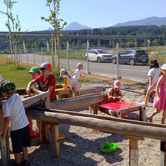 Eine Gruppe von Kindern spielt in einem Spielplatz mit Schotterboden und Holzstrukturen. Dahinter steht ein Metallzaun mit einer Straße und zwei geparkten Autos. Bäume und Berge sind in der Ferne.