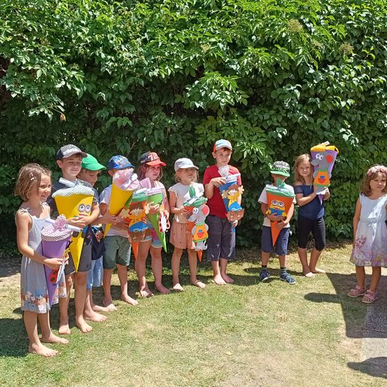 Eine Gruppe von Kindern in Sommerkleidung steht in einem Außenbereich und hält bunte Eistüten, mit einer grünen Hecke im Hintergrund.
