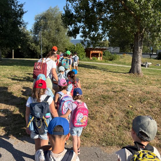 Eine Gruppe von Kindern geht in einer Reihe, mit Rucksäcken, auf einem Pfad in einem Park, mit einem Baum auf der rechten Seite.