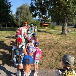 Eine Gruppe von Kindern geht in einer Reihe, mit Rucksäcken, auf einem Pfad in einem Park, mit einem Baum auf der rechten Seite.