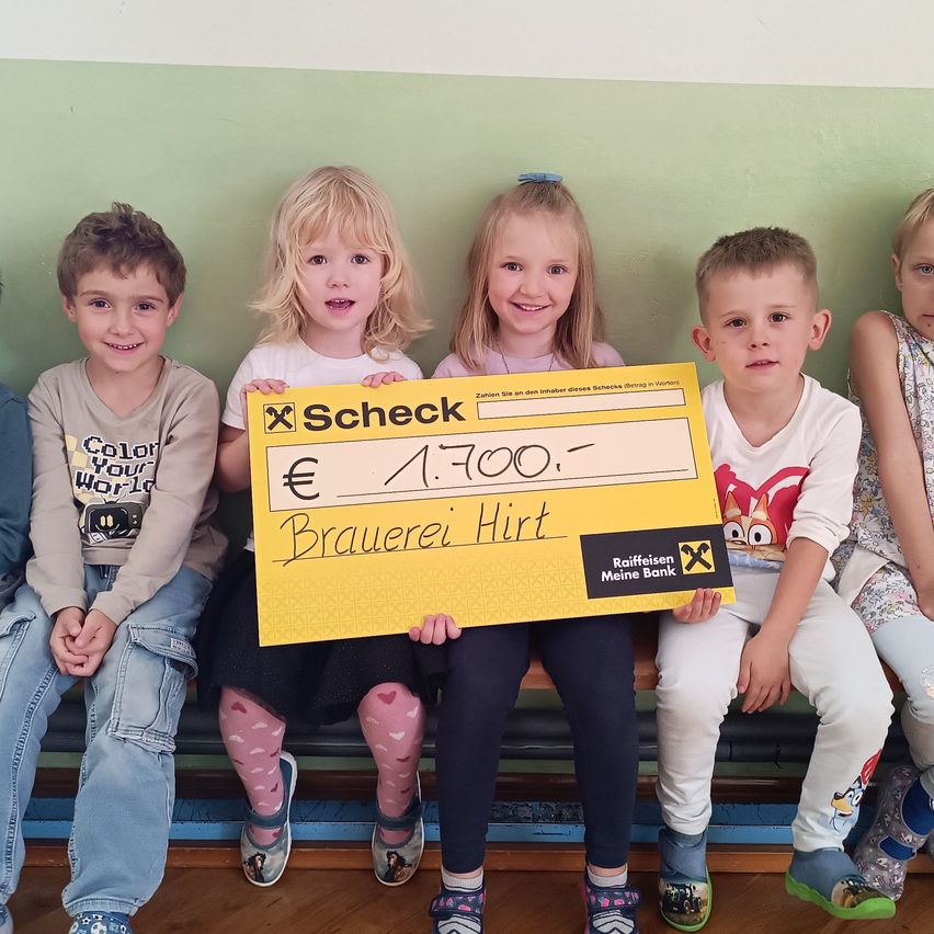 Fünf junge Kinder sitzen lächelnd auf einer Bank. Ein Mädchen in der Mitte hält einen großen gelben Scheck über 1.700 Euro, unterzeichnet von Brauerei Hirt. Der Scheck stammt von Raiffeisen Meine Bank. Die Kinder tragen Freizeitkleidung, und der Hintergrund ist eine grüne Wand.