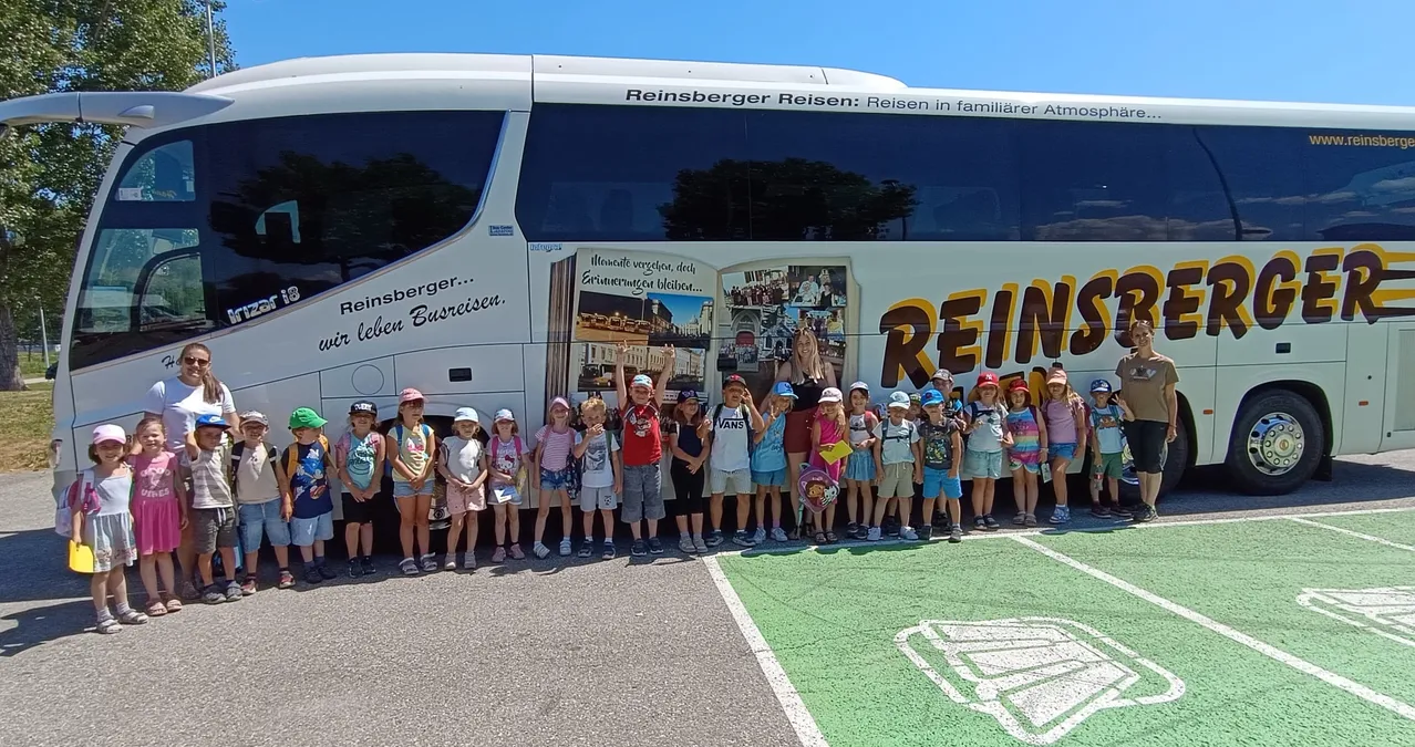 Eine Gruppe von Kindern in passenden Outfits steht vor einem weißen Bus mit dem Text 'Reinsberg Reisen' und einer Collage an der Seite. Sie posieren für ein Foto.