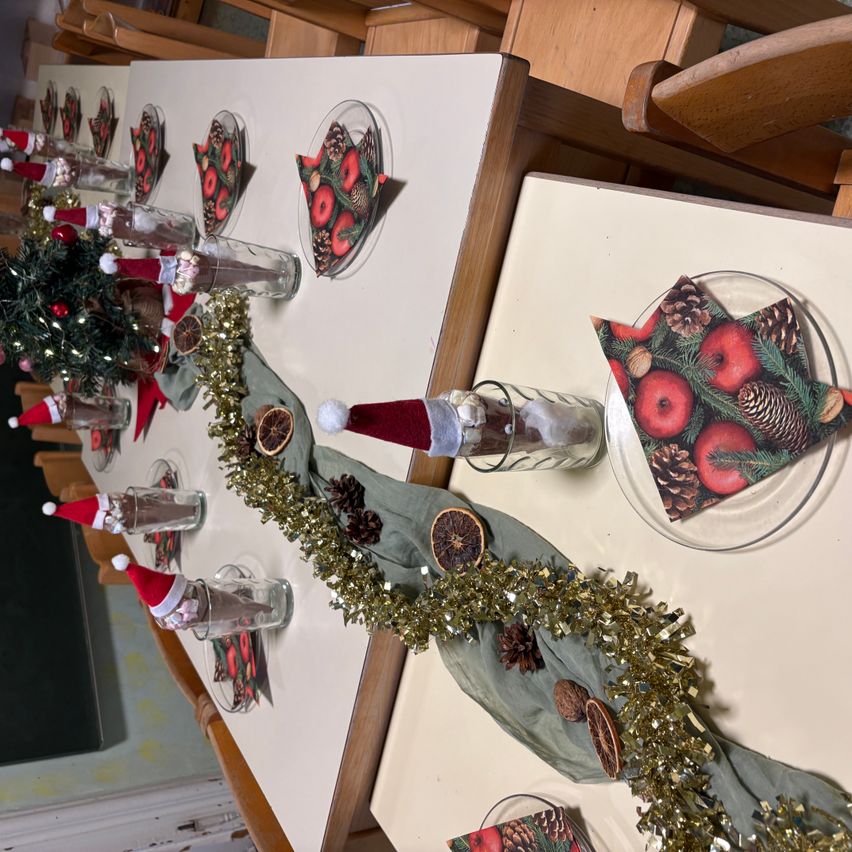 Eine festliche Tischdekoration mit Weihnachtsdekorationen, einschließlich Tannenzapfen, Ornamenten und Glitzerband. Jede Tischplatzierung hat ein Serviettentuch, ein Glas und einen Weihnachtsmannhut.