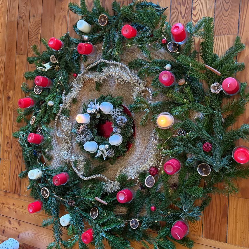 Ein runder Kranz mit grünen Zweigen und roten Kerzen liegt auf einem Holzboden. Der Kranz ist mit silbernen und weißen Ornamenten geschmückt, und einige Kerzen sind angezündet. Es gibt auch getrocknete Früchte und Tannenzapfen im Kranz.