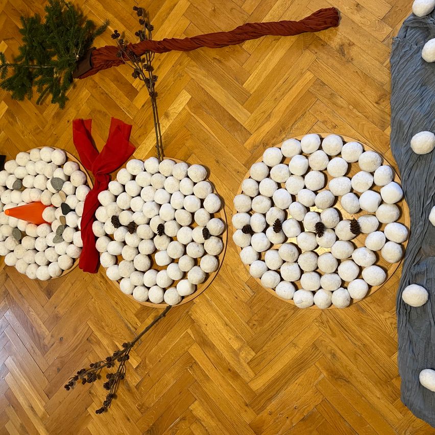 Drei Schneemänner aus weißen Kugeln sind auf einem Holzboden angeordnet. Jeder Schneemann hat ein rotes Band, eine Karottennase und Tannenzapfen. Der Hintergrund ist unscharf, wobei ein kleiner Baum und ein grauer Stoff sichtbar sind.