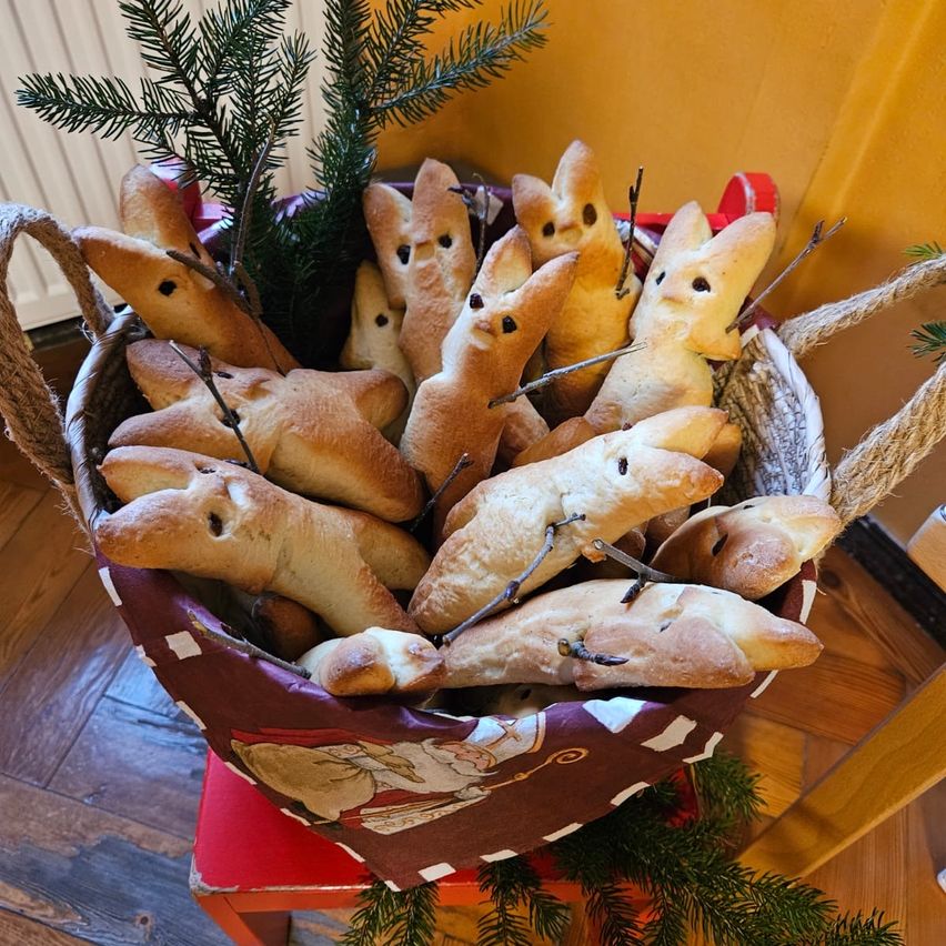 Ein Korb mit festlichen Brötchen in Form von Hasen mit Augen und Zweigen, auf einem Holzboden mit gelben Wänden und Weihnachtsdekorationen.