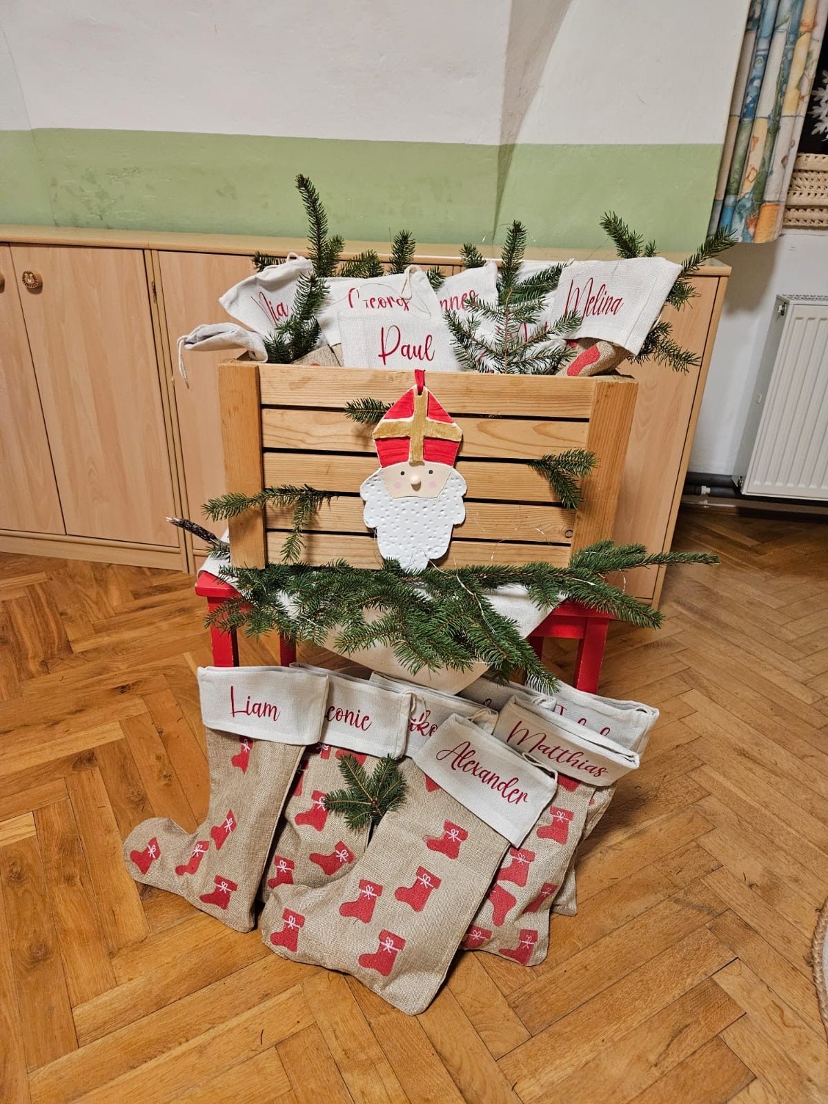 Ein hölzerner Kasten mit Tannenzweigen und dem Weihnachtsmann darauf. Mehrere personalisierte Socken sind darunter angeordnet, jede mit einem Namen wie Liam, Connie, Alexander und Matthias. Die Umgebung besteht aus Holzschränken und einem Boden.