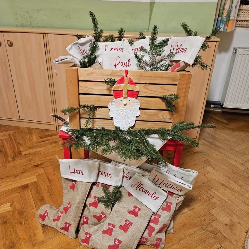 Ein hölzerner Kasten mit Tannenzweigen und dem Weihnachtsmann darauf. Mehrere personalisierte Socken sind darunter angeordnet, jede mit einem Namen wie Liam, Connie, Alexander und Matthias. Die Umgebung besteht aus Holzschränken und einem Boden.