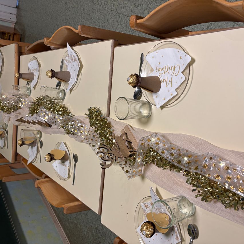 Ein Tisch, der für ein festliches Event vorbereitet ist, mit goldenem Tinsel, Papiertüten, Schokoladen und Besteck. Jede Tischdecke hat ein gefaltetes Papiertuch, einen Teller und ein Glas.