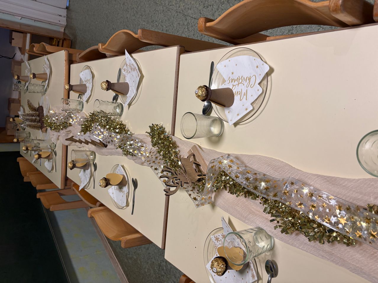 Ein Tisch, der für ein festliches Event vorbereitet ist, mit goldenem Tinsel, Papiertüten, Schokoladen und Besteck. Jede Tischdecke hat ein gefaltetes Papiertuch, einen Teller und ein Glas.