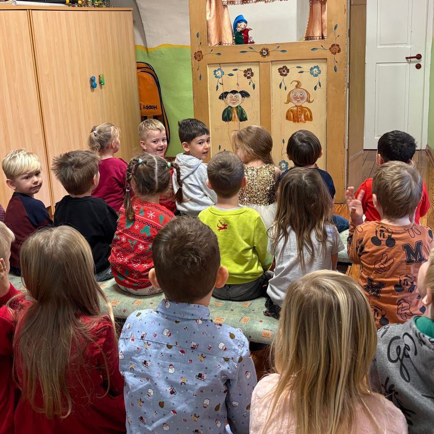 Kinder sitzen auf einer Bank in einem Raum und schauen sich eine Puppenshow an. Hinter ihnen steht eine Holztür mit gemalten Figuren und links befindet sich ein Holzschrank.