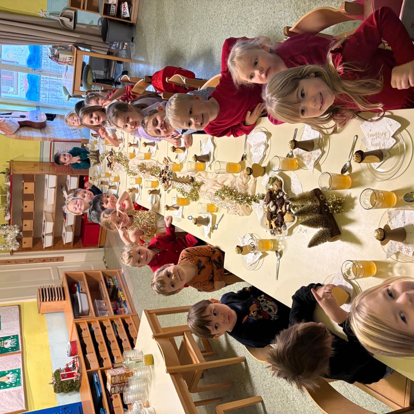 Eine Gruppe von Kindern sitzt um einen Tisch mit Essen und Getränken. Einige von ihnen tragen rote Hemden. Der Tisch ist mit Tellern, Gläsern und Servietten gedeckt. Es gibt auch Dekorationen auf dem Tisch. Dahinter ist eine Wand mit einem Weihnachtsbaum und einem Fenster.